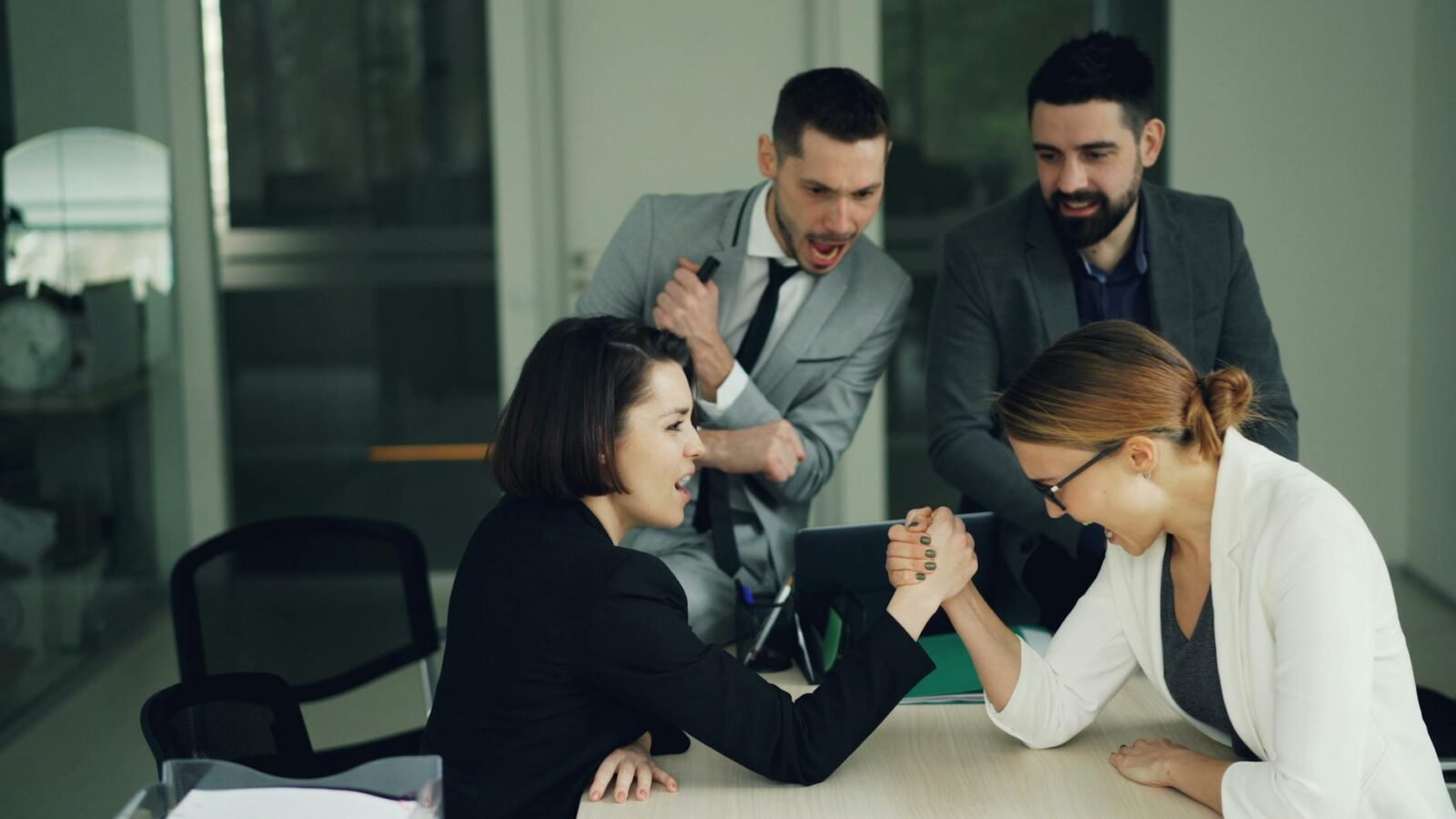 Zwei Kolleginnen liefern sich in einem Büro ein Armdrücken, während männliche Kollegen sie anfeuern.