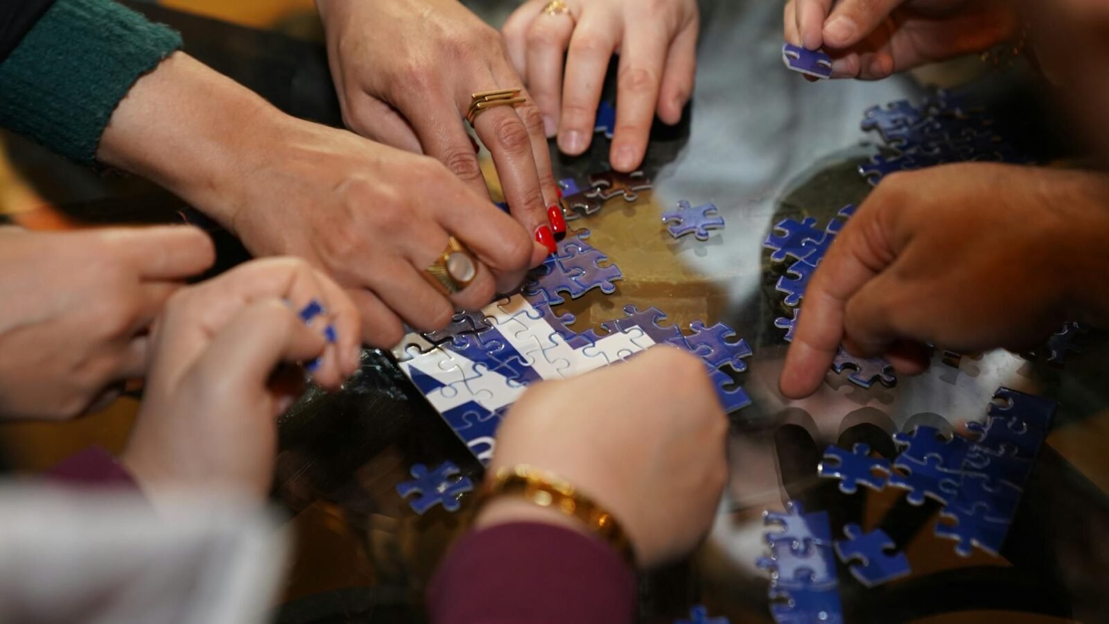 Eine bunt zusammengewürfelte Gruppe von Menschen setzt gemeinsam ein Puzzle an einem Tisch in einem Innenraum zusammen.